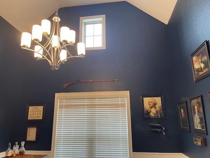 High-ceilinged room with navy walls, chandelier, window, gun, framed portraits, and window blinds.