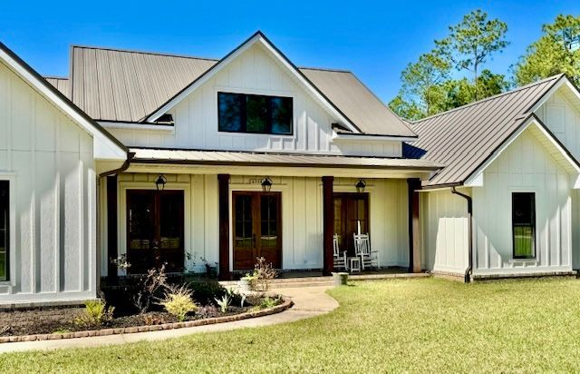 White farmhouse with metal roof, dark brown doors, front porch, and green lawn.