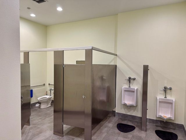 Public restroom with stainless steel stalls and urinals, light beige walls, gray floor.