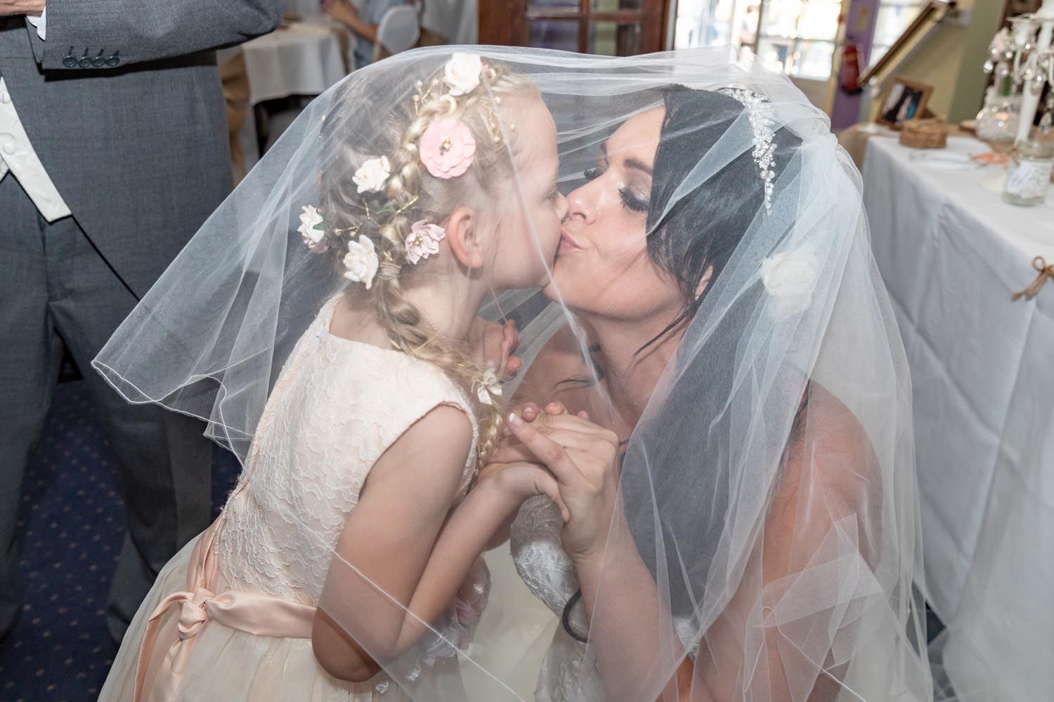 A bride and a flower girl are kissing under a veil.