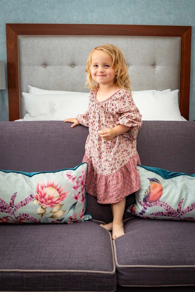 A little girl is standing on a purple couch next to a bed.