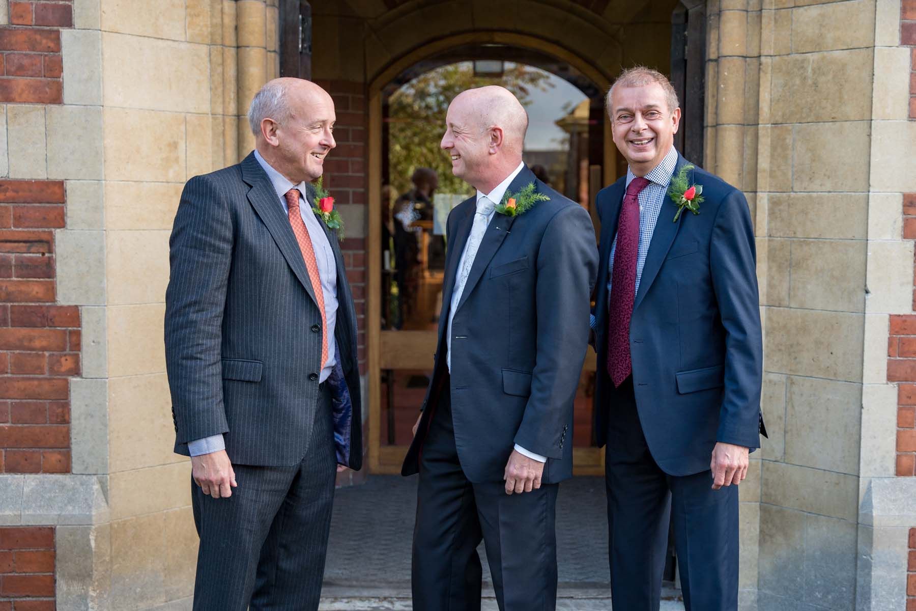 Three men in suits and ties are standing in front of a brick building.