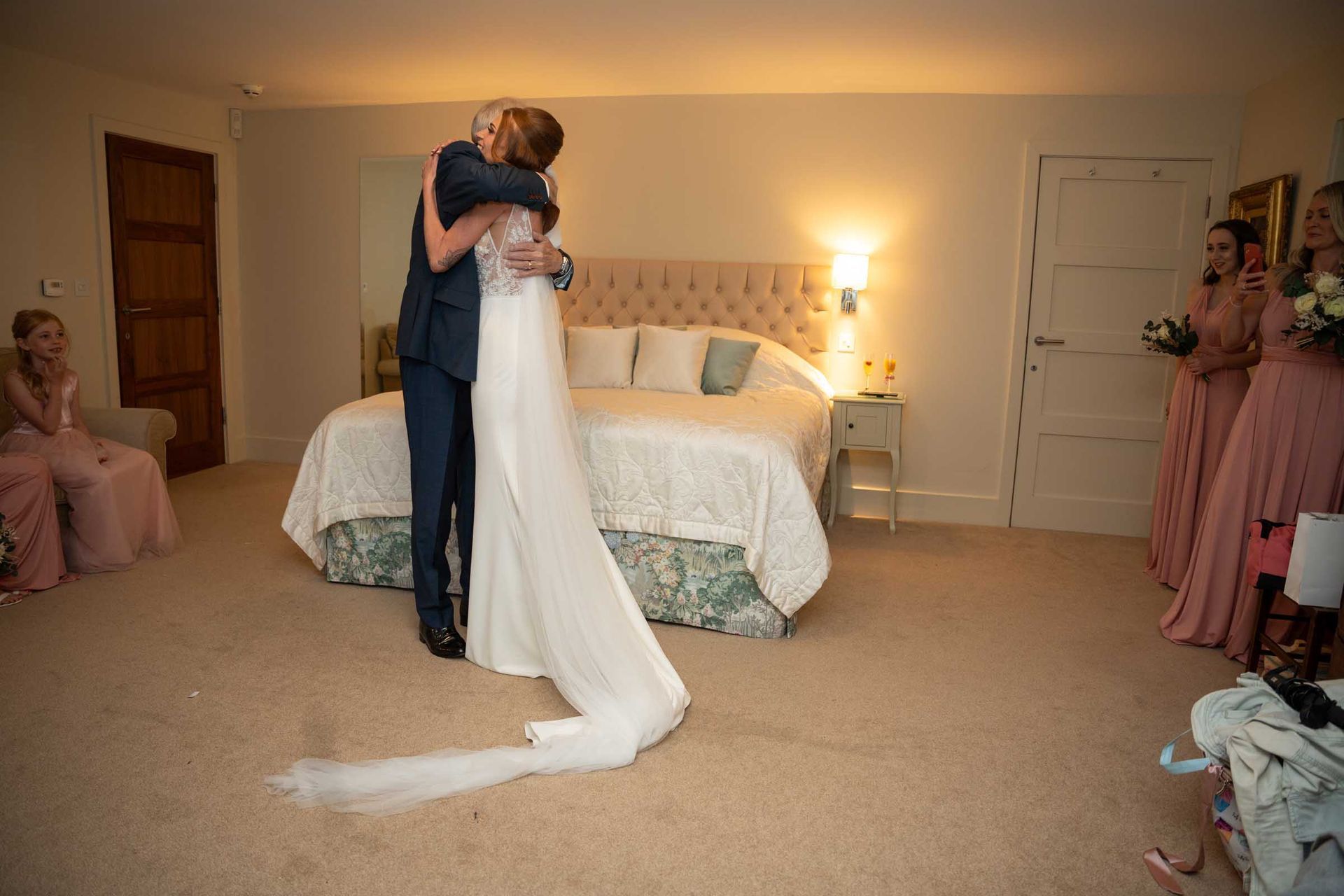 A bride and groom are hugging in a bedroom while their bridesmaids watch.