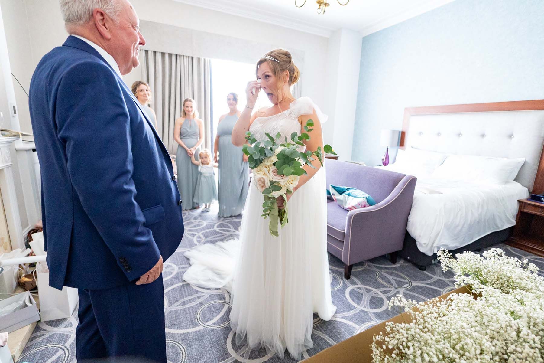 A bride is crying while standing next to her father in a hotel room.