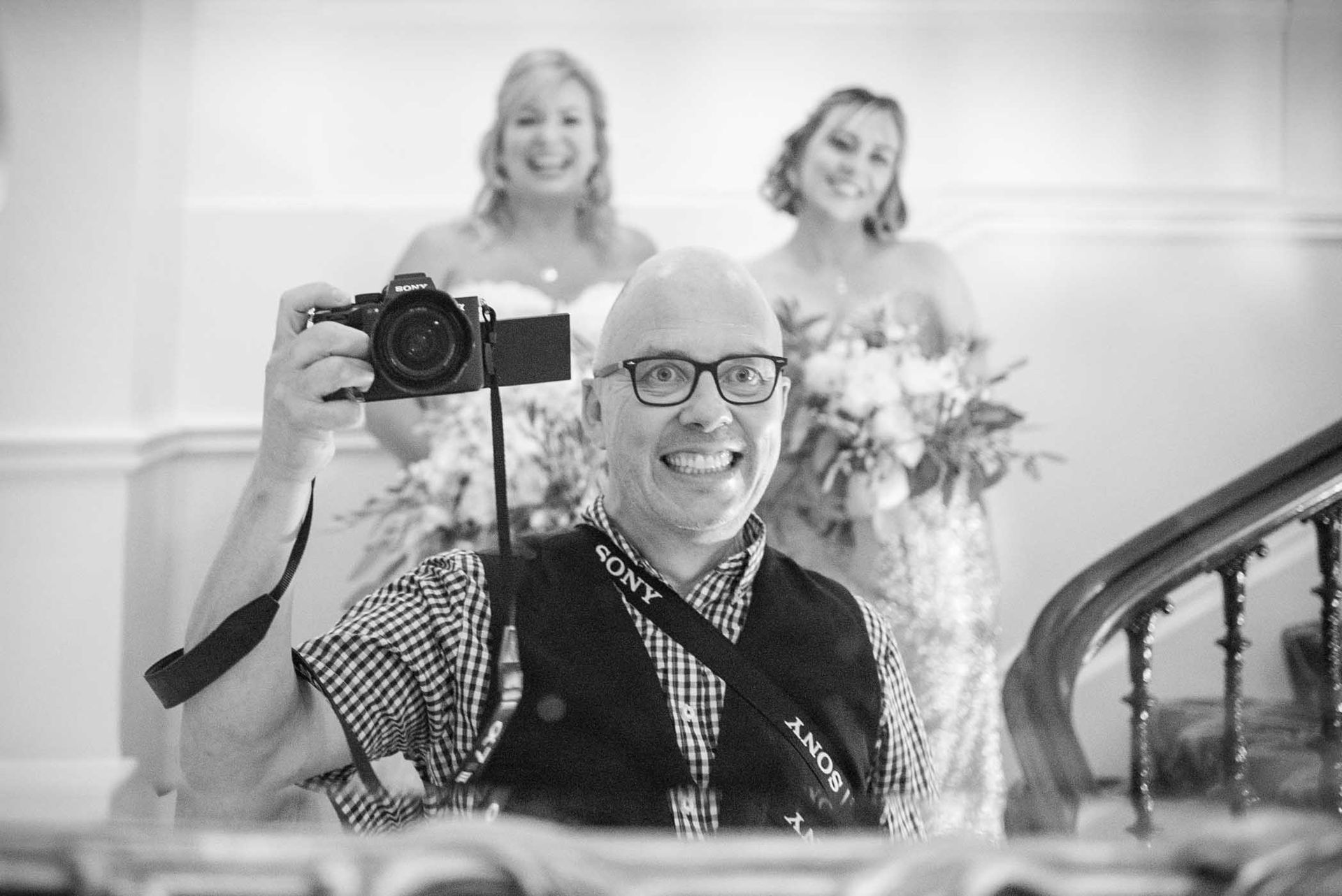 A man is taking a picture of himself in a mirror with two women in the background.