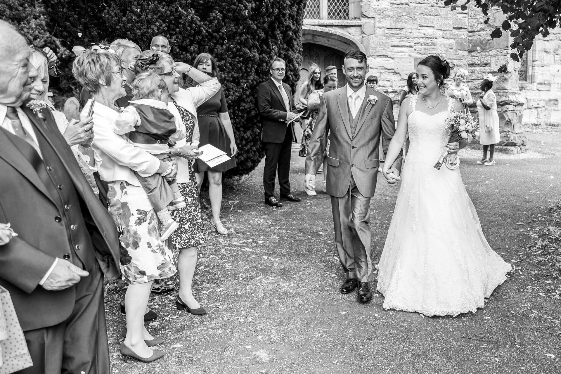 A black and white photo of a bride and groom walking down a path with their wedding guests.