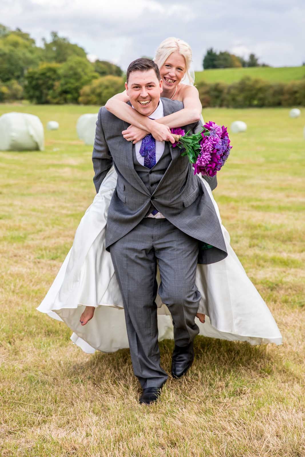 A man is carrying a bride on his back in a field.