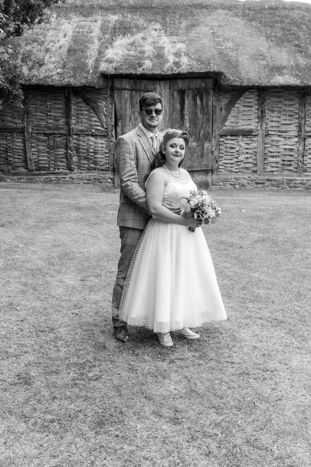 A bride and groom are posing for a black and white photo in front of a thatched barn.