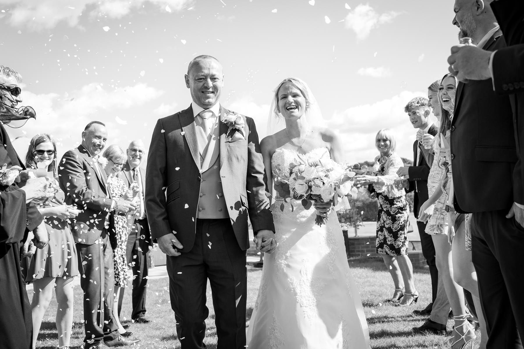 A black and white photo of a bride and groom walking through confetti.