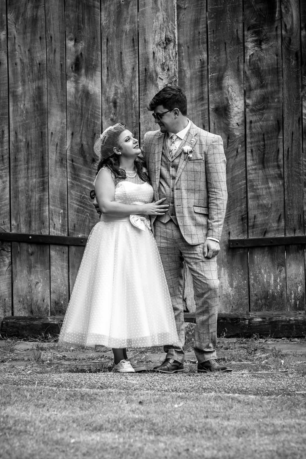 A black and white photo of a bride and groom standing next to each other in front of a wooden fence.