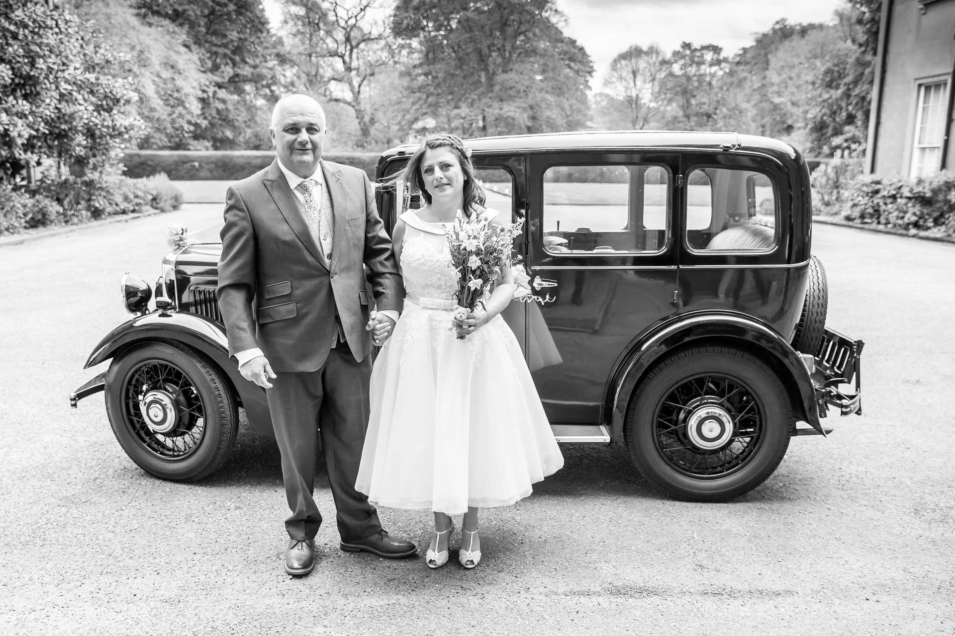 A black and white photo of a bride and groom standing next to an old car.