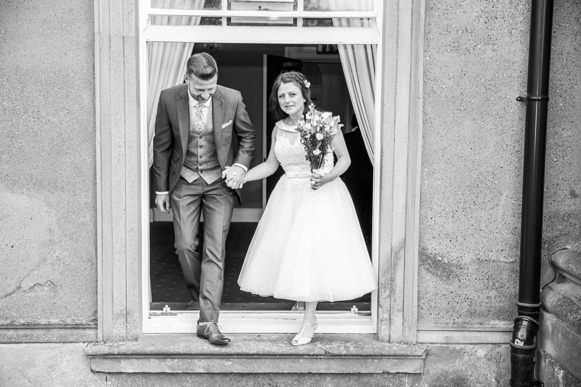 A bride and groom are walking out of a window holding hands.