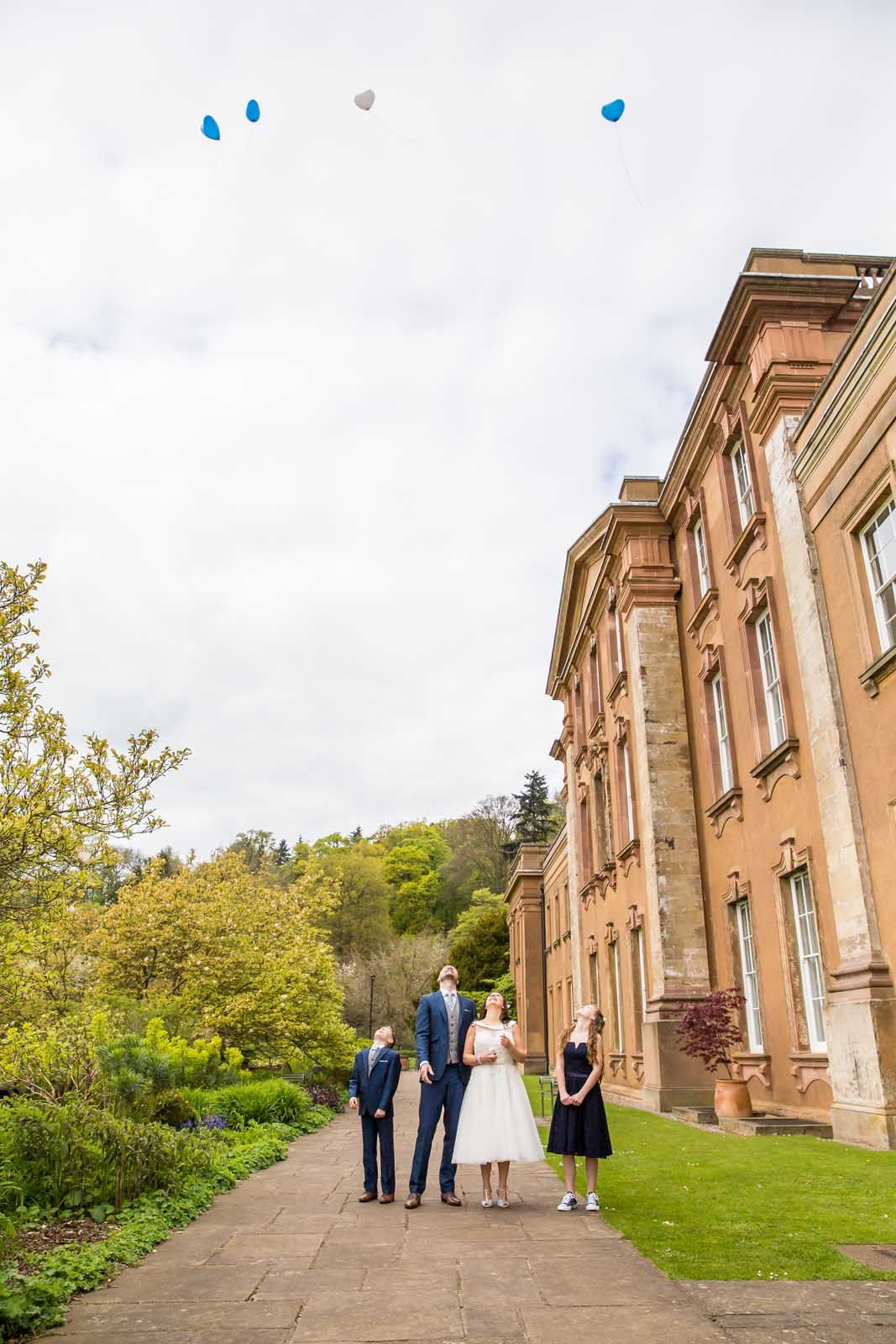 A bride and groom are standing in front of a large building with balloons flying in the air.