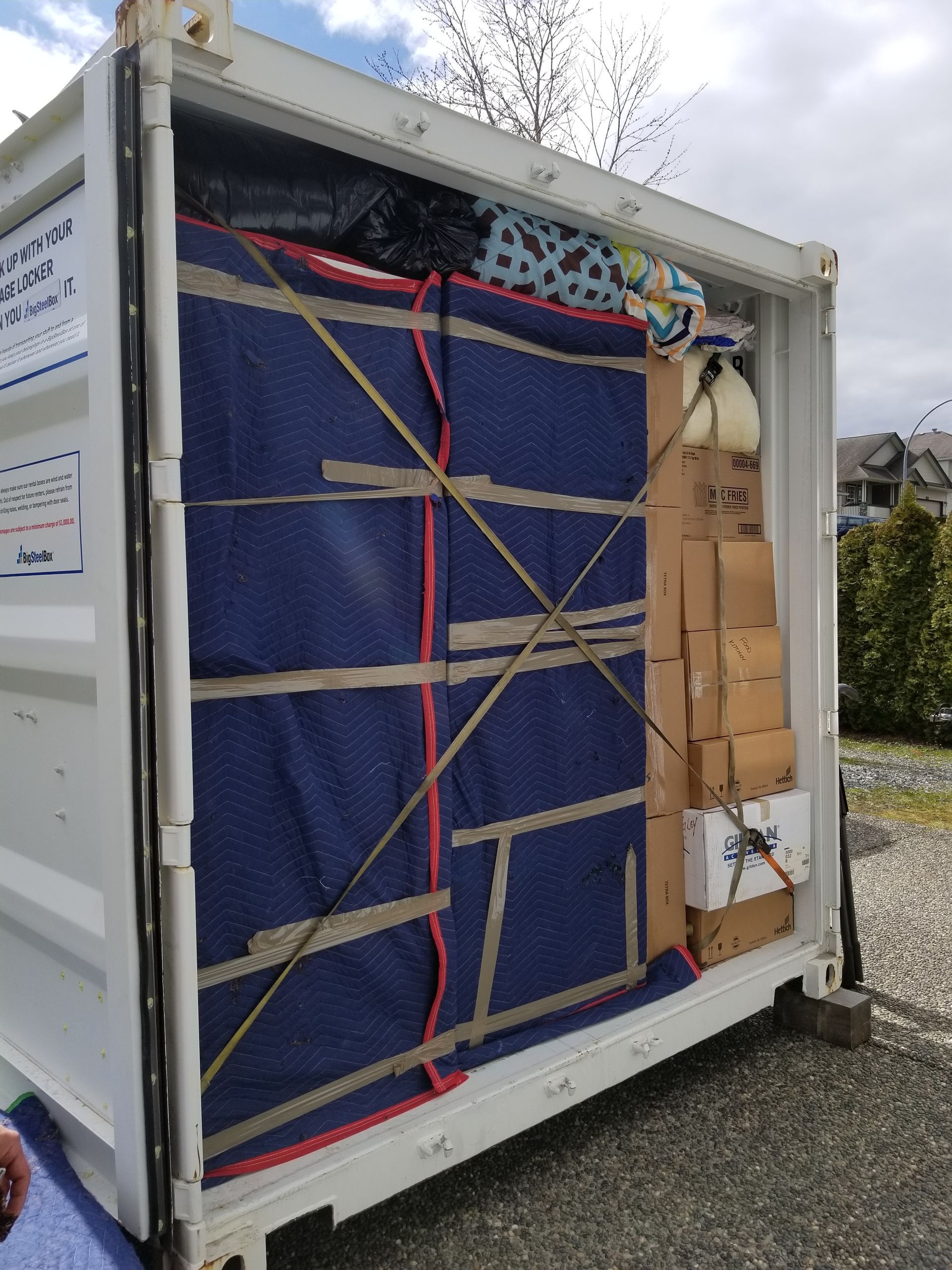 Shipping container packed with furniture pads, boxes, and other items, secured with straps. Container is open with items visible.