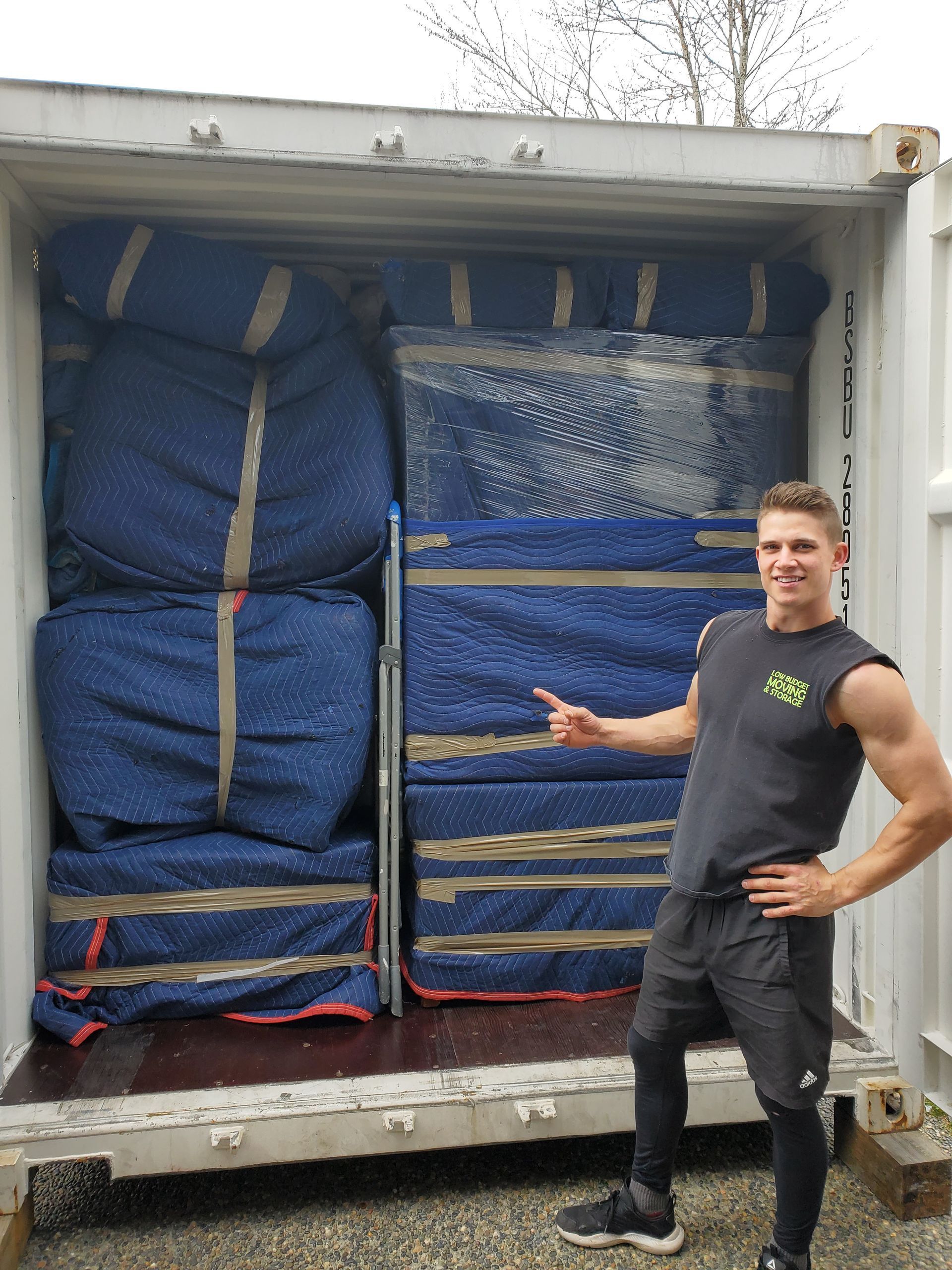 A man points to a shipping container packed with blue-wrapped items. He's standing in the container, smiling.