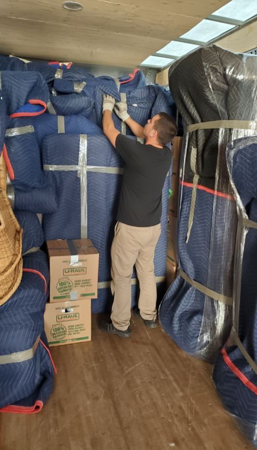 Man securing packed furniture with tape inside a moving truck. Boxes and blue padded wraps fill the space.