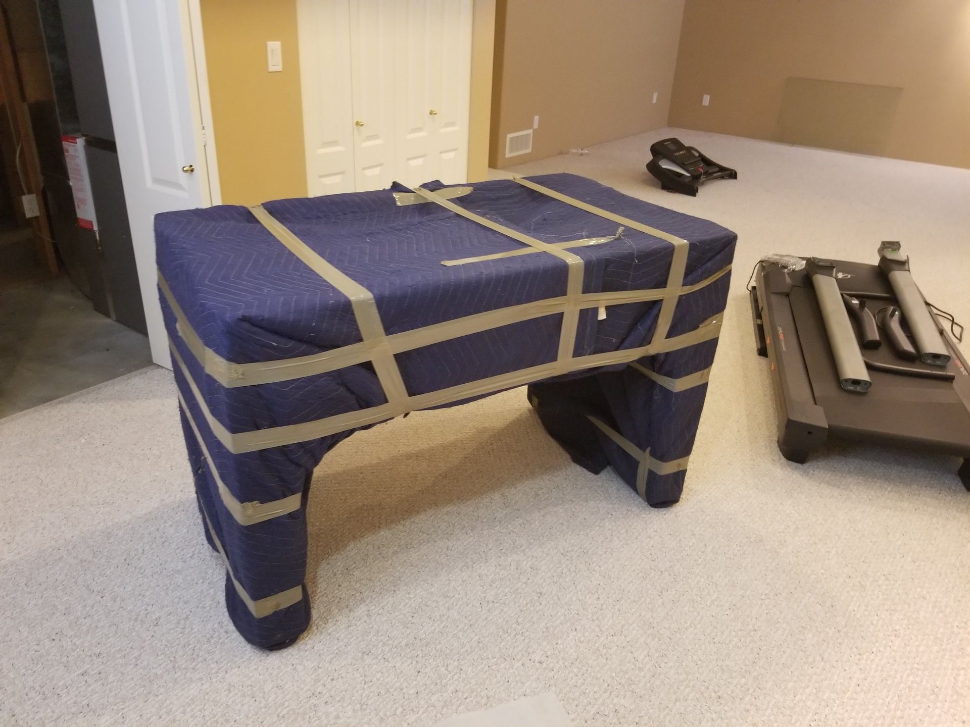 A table wrapped in blue moving blankets and tape, next to a folded treadmill in a room with beige walls.