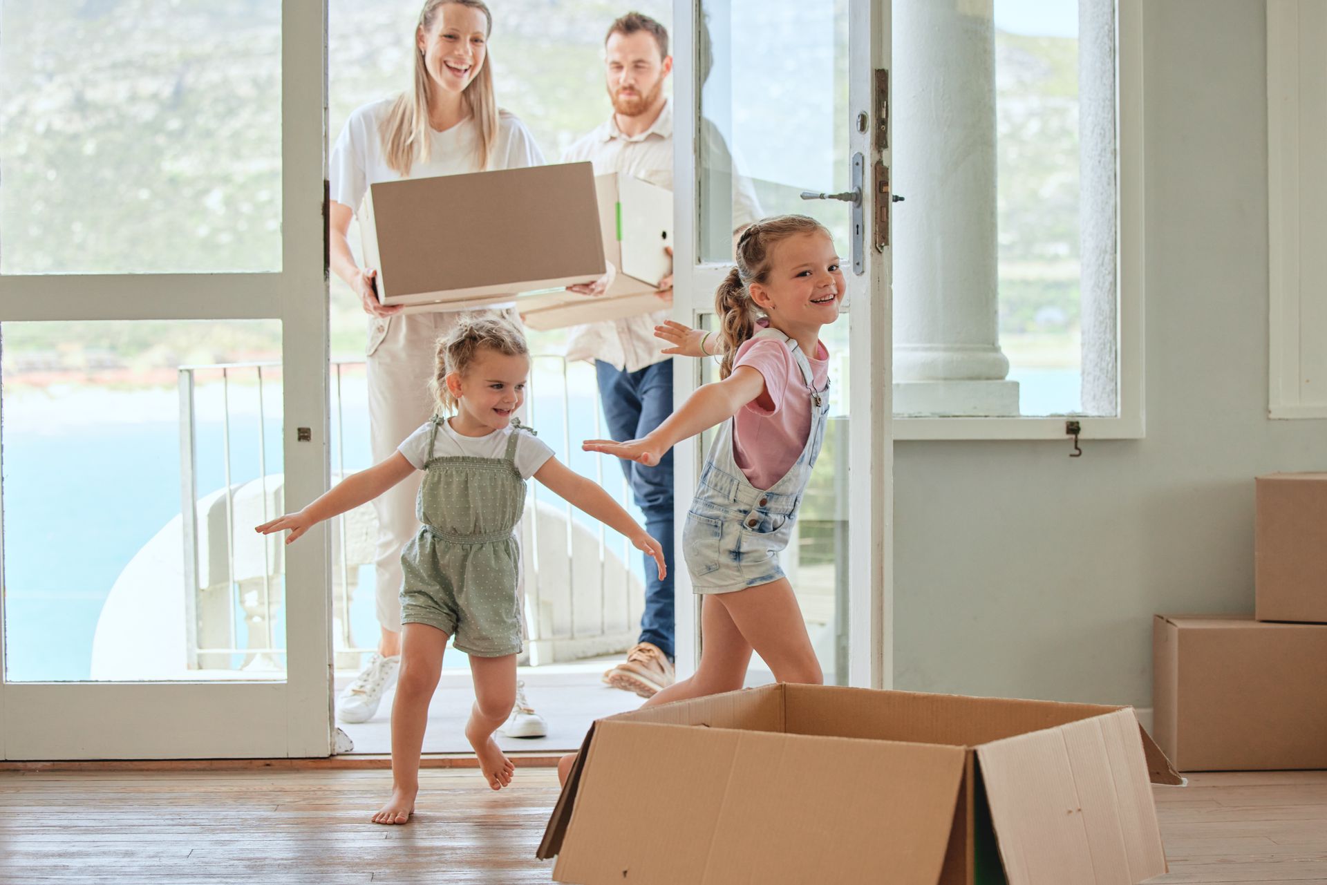 Family of four moving into a new home; children running ahead, parents carrying boxes, sunny doorway.