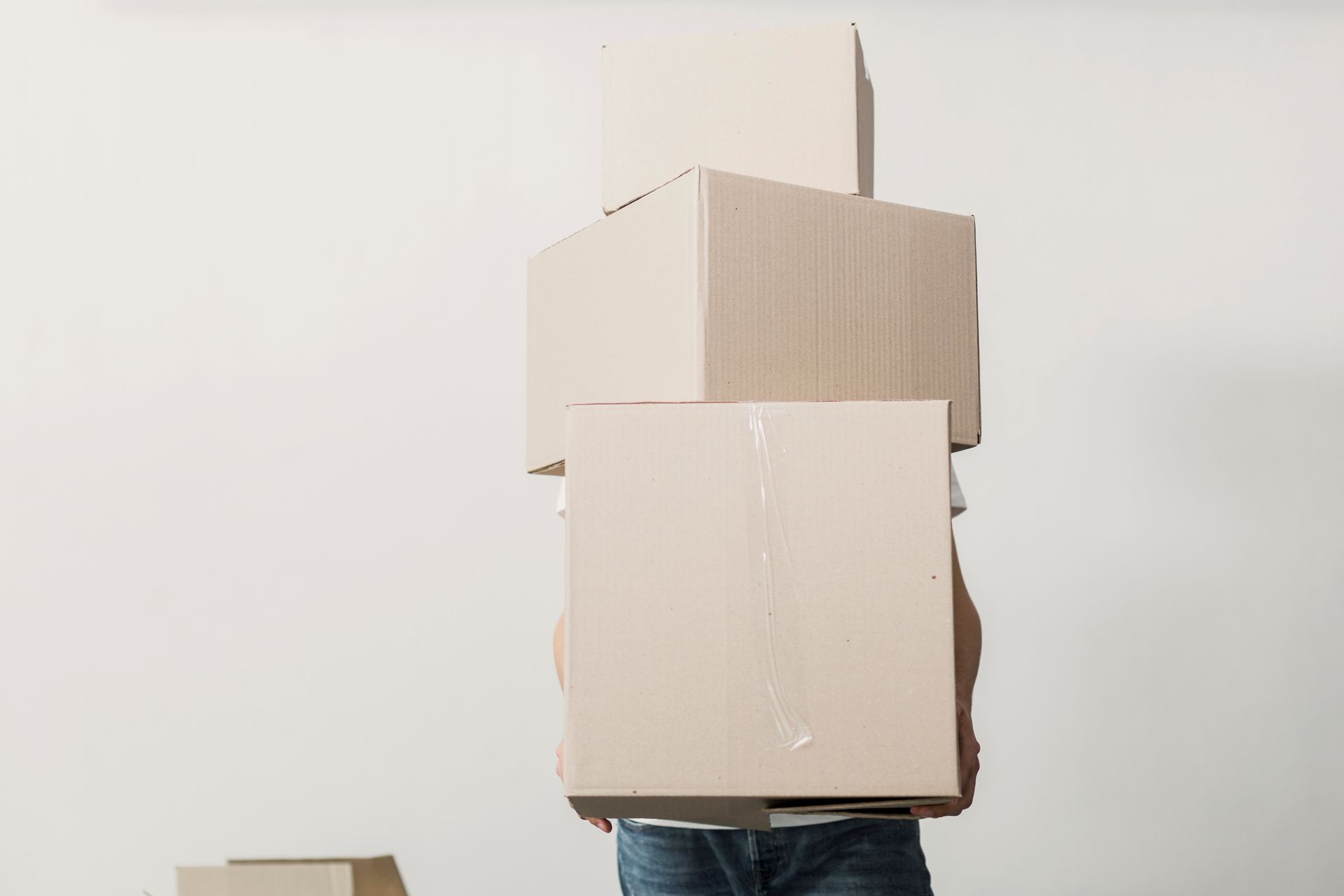 Person carrying a stack of beige cardboard boxes in front of a light wall.