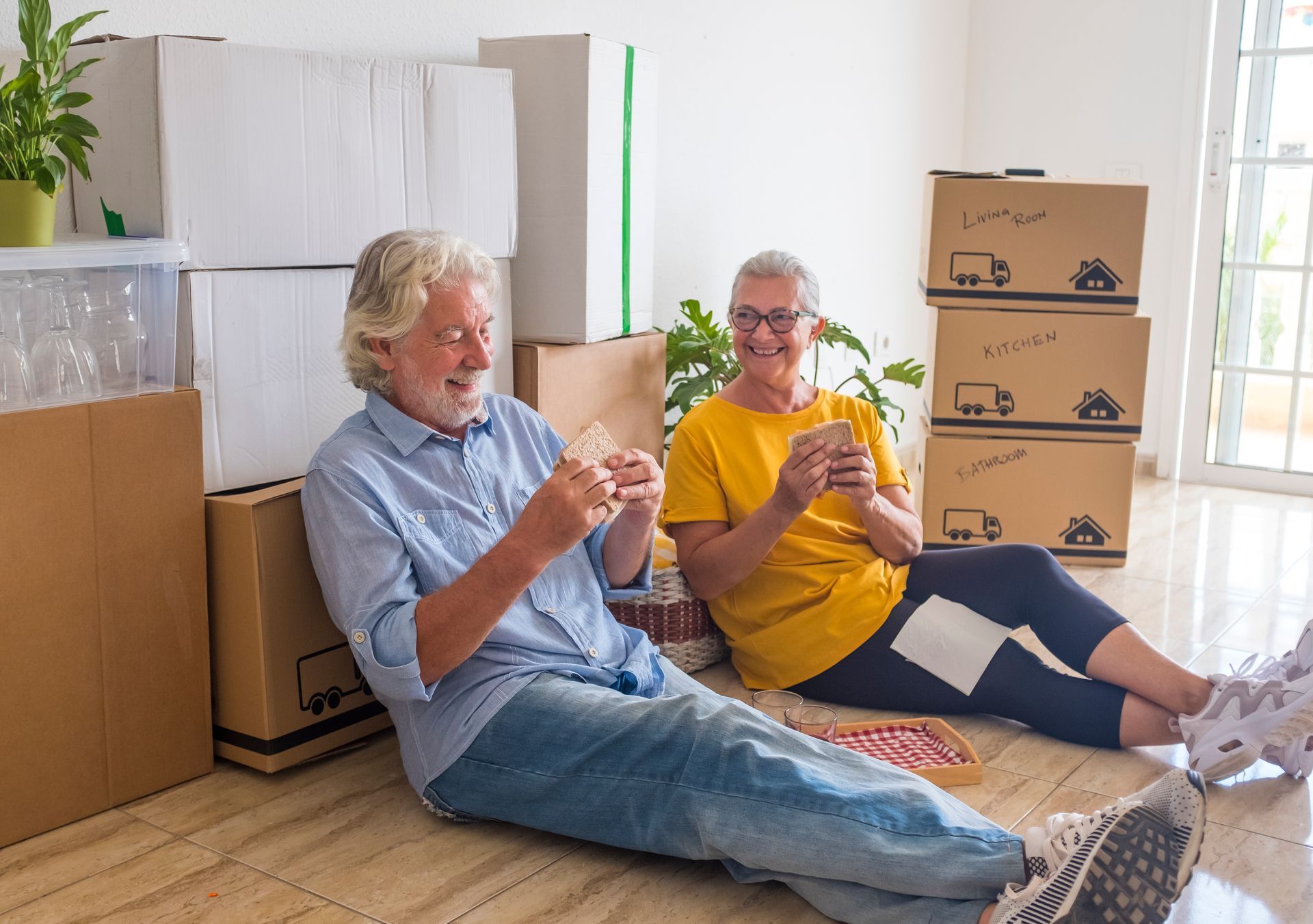 An elderly couple is sitting on the floor in front of moving boxes.