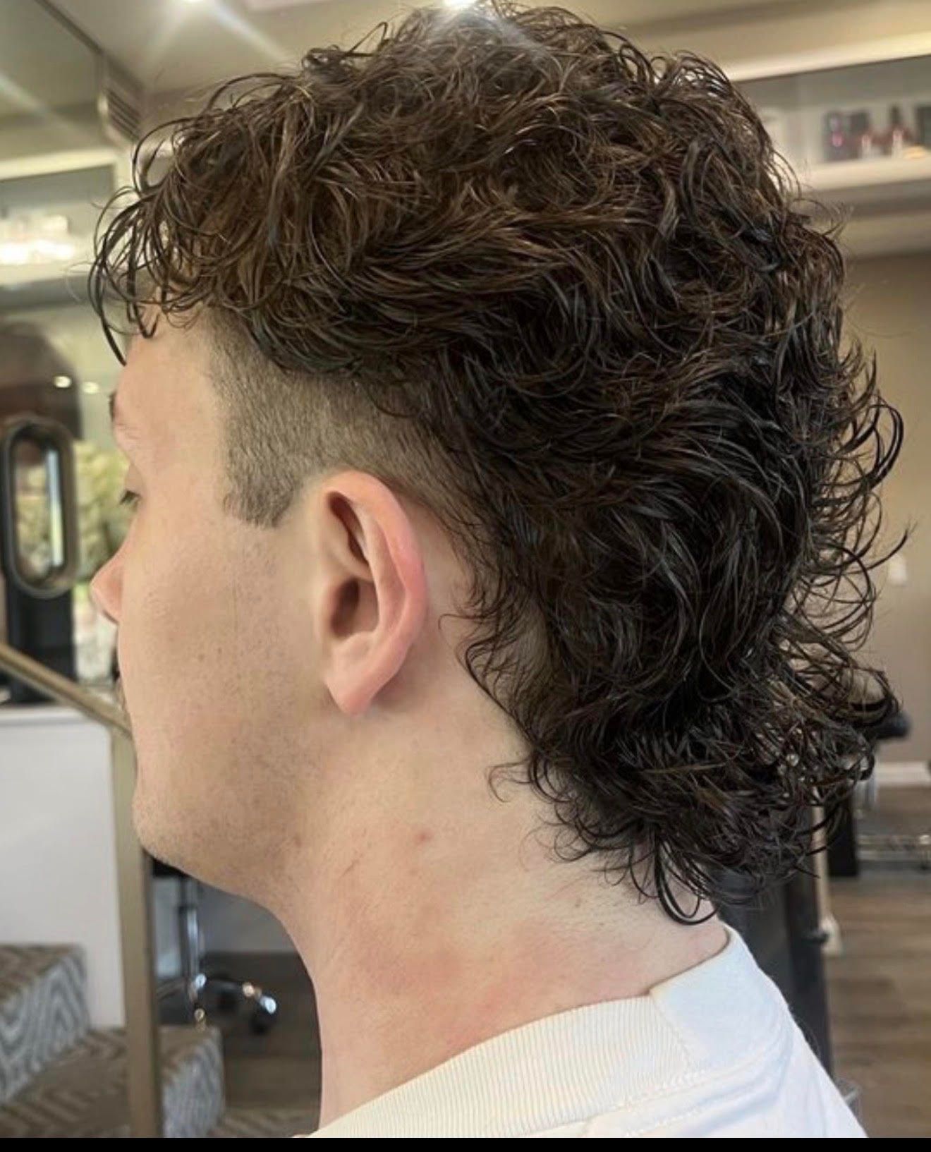 Side view of a person with curly dark hair and a mullet-style haircut in a barbershop.