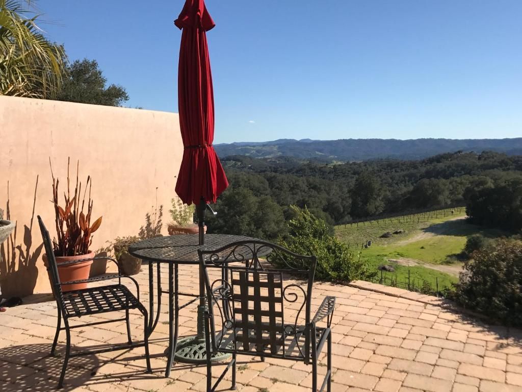 Patio with black metal furniture, red umbrella, and a view of a valley and vineyard on a sunny day.