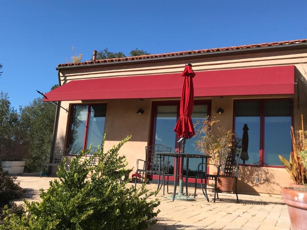 Red awning shades a patio with a table, chairs, and a red umbrella; tan house with windows and a blue sky.