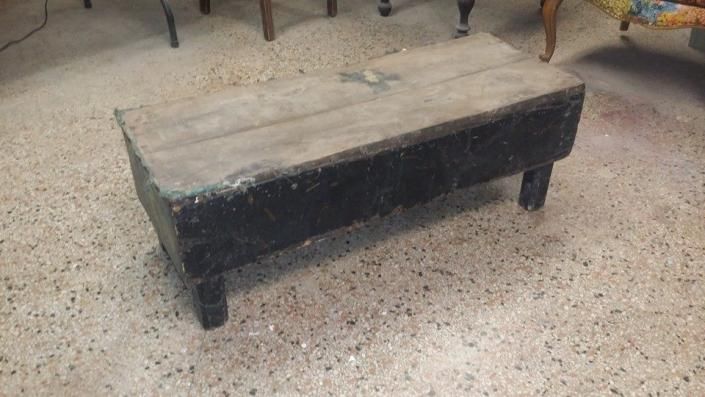 Worn black upholstered bench on short legs with a beige top, sitting on a speckled floor.