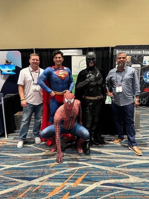 Group posing with Spider-Man, Superman, and Batman at an event, near a booth.