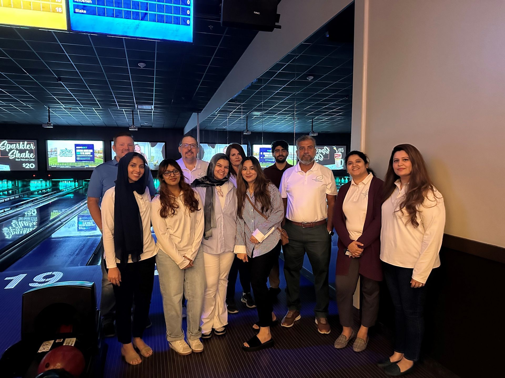 Group of people at a bowling alley. Several smiling and posing together, in front of the bowling lanes.