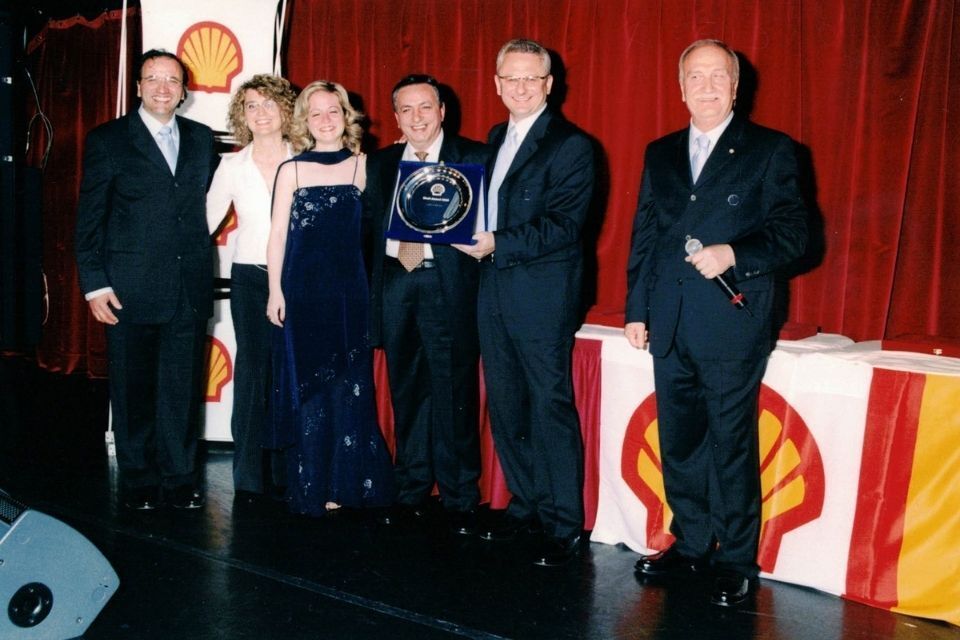 Persone sul palco con in mano un trofeo durante un evento Shell, sullo sfondo una tenda rossa e uno striscione con il logo Shell.