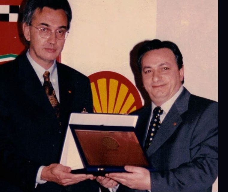 Due uomini in giacca e cravatta tengono in mano un premio. Uno di loro porge una targa all'altro, davanti al logo della Shell.