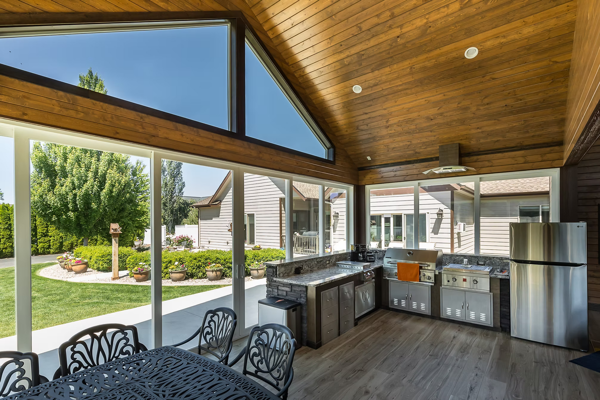 Outdoor kitchen with stainless steel appliances and seating, overlooking a grassy yard and garden.