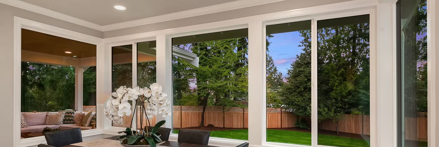 View through large windows of a yard with trees, lawn, and a fence. Bright interior.