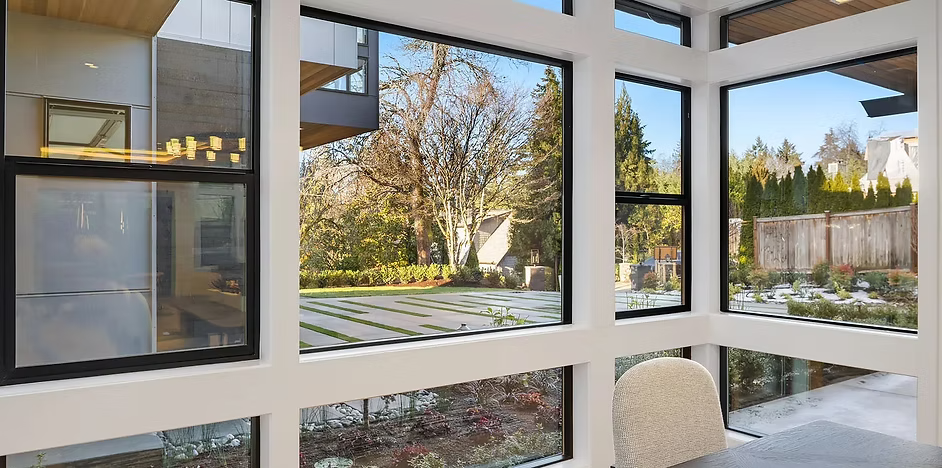 Modern corner room with large windows framing a yard, trees, and a house.