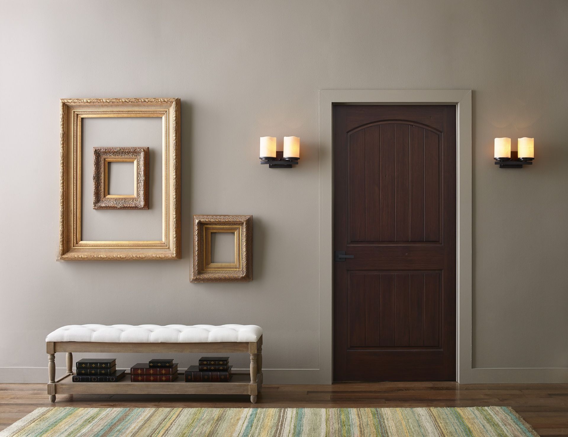 Hallway with dark wood door, empty gold frames, bench, sconces, and a rug.