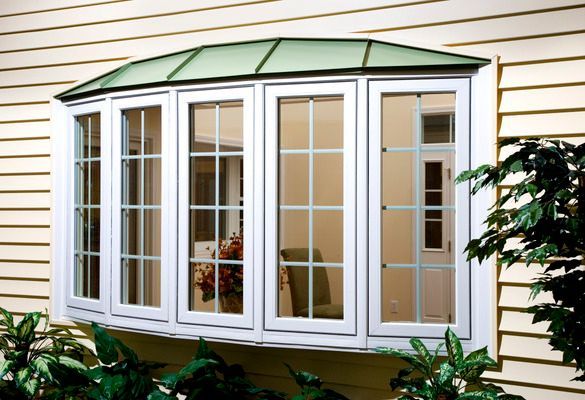 Bay window with white frames and grid pattern on a yellow house. Green roof and foliage in foreground.