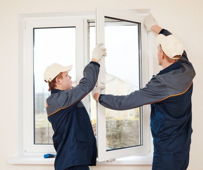 Two workers installing a white window, one holding the frame, the other adjusting it indoors.