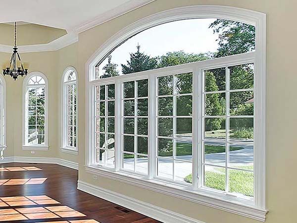 Large arched window with grid panes in a beige room, overlooking a green yard and trees.