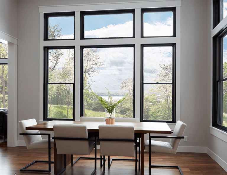Dining room with large windows, view of nature, wooden table, white chairs, and vase.