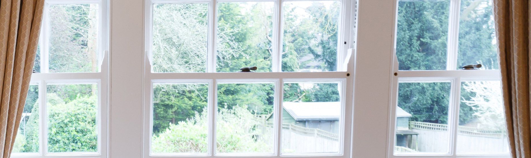 Three white-framed windows with greenery visible through the panes, flanked by tan curtains.