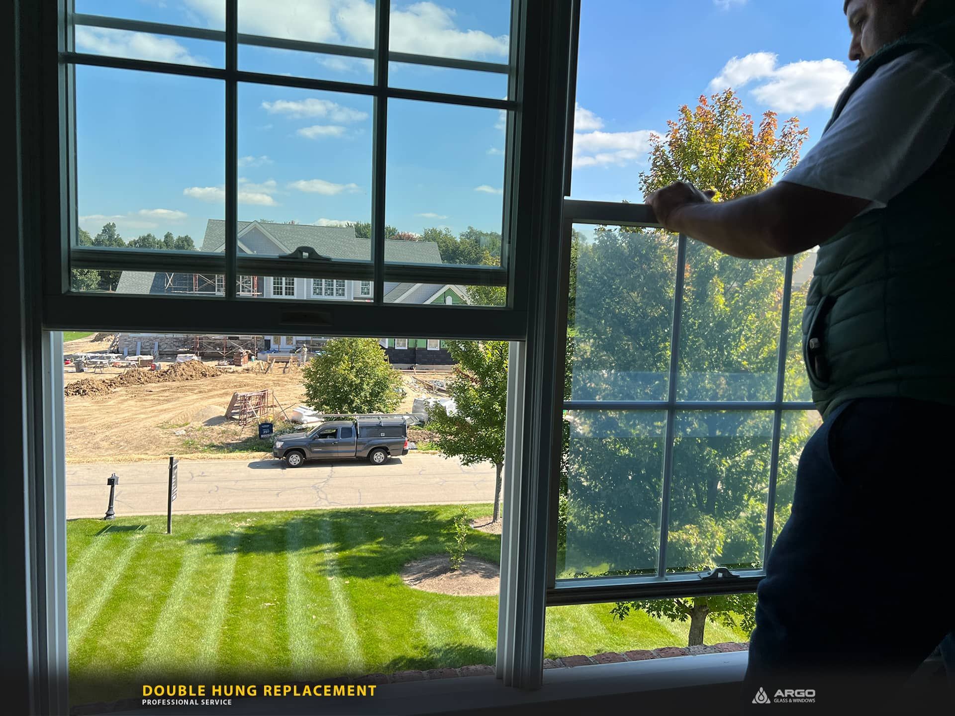 Person replacing a double-pane window. Shows the interior view, a lawn, and construction beyond. Blue sky.