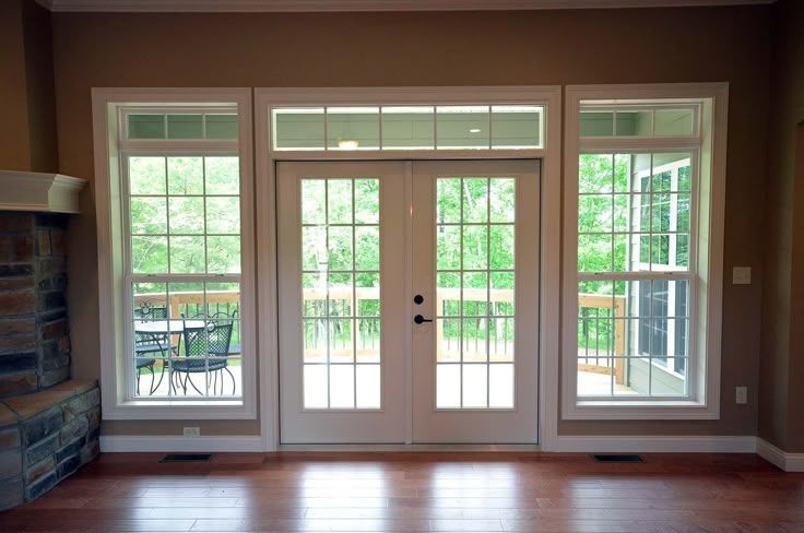 French doors and windows overlooking a deck and trees; white trim, brown walls, wood floor.