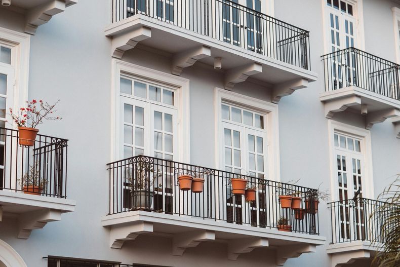 Light blue building with white-framed windows and black wrought-iron balconies holding potted plants.