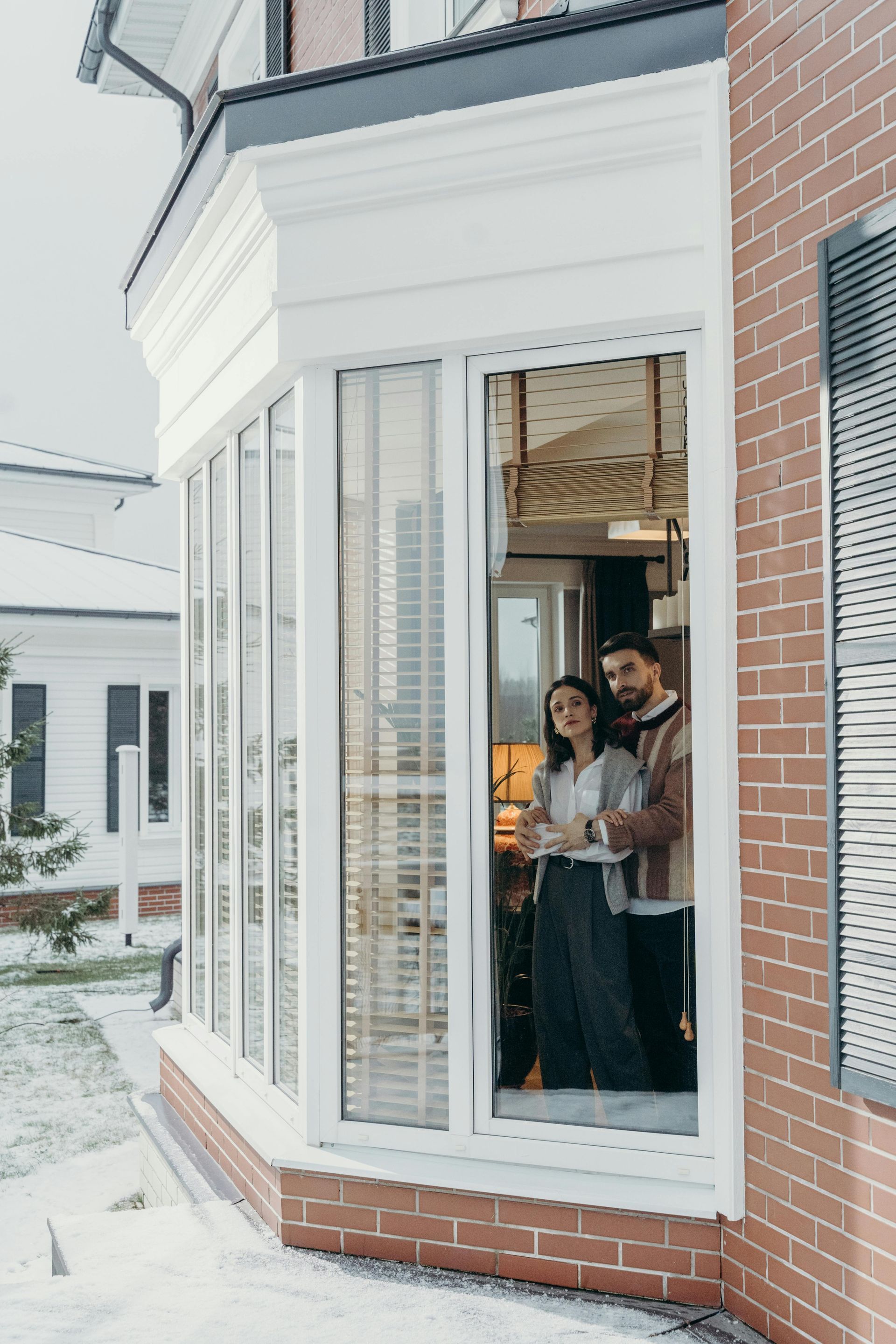 Couple standing inside a bay window of a brick house, looking out at a snowy yard.