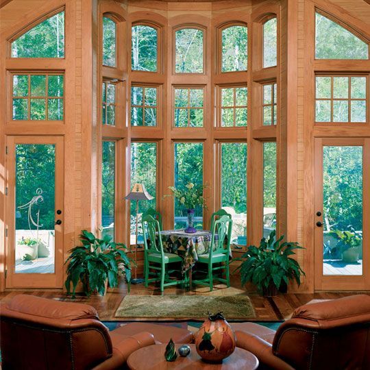 Sunroom with large wooden windows, green dining set, and brown leather armchairs. Lush green outdoor view.