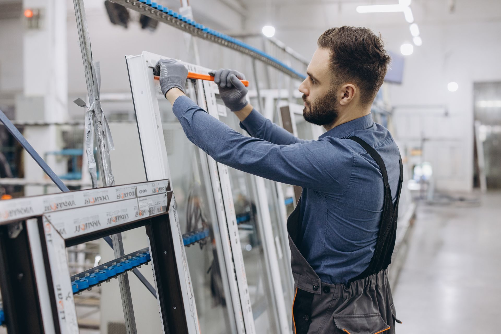Man working on window frame in a factory, wearing gloves and overalls.