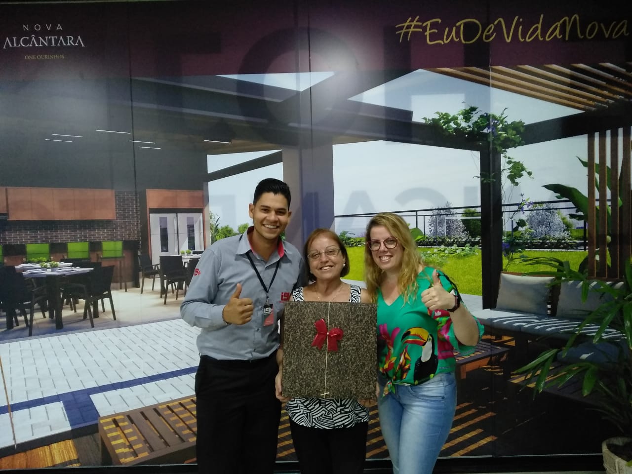 Três pessoas posam para foto em frente a uma placa que diz eu da vida nova