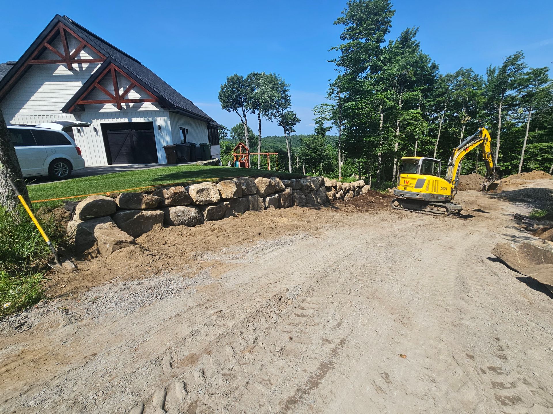 Une excavatrice jaune travaille sur un chemin de terre devant une maison.