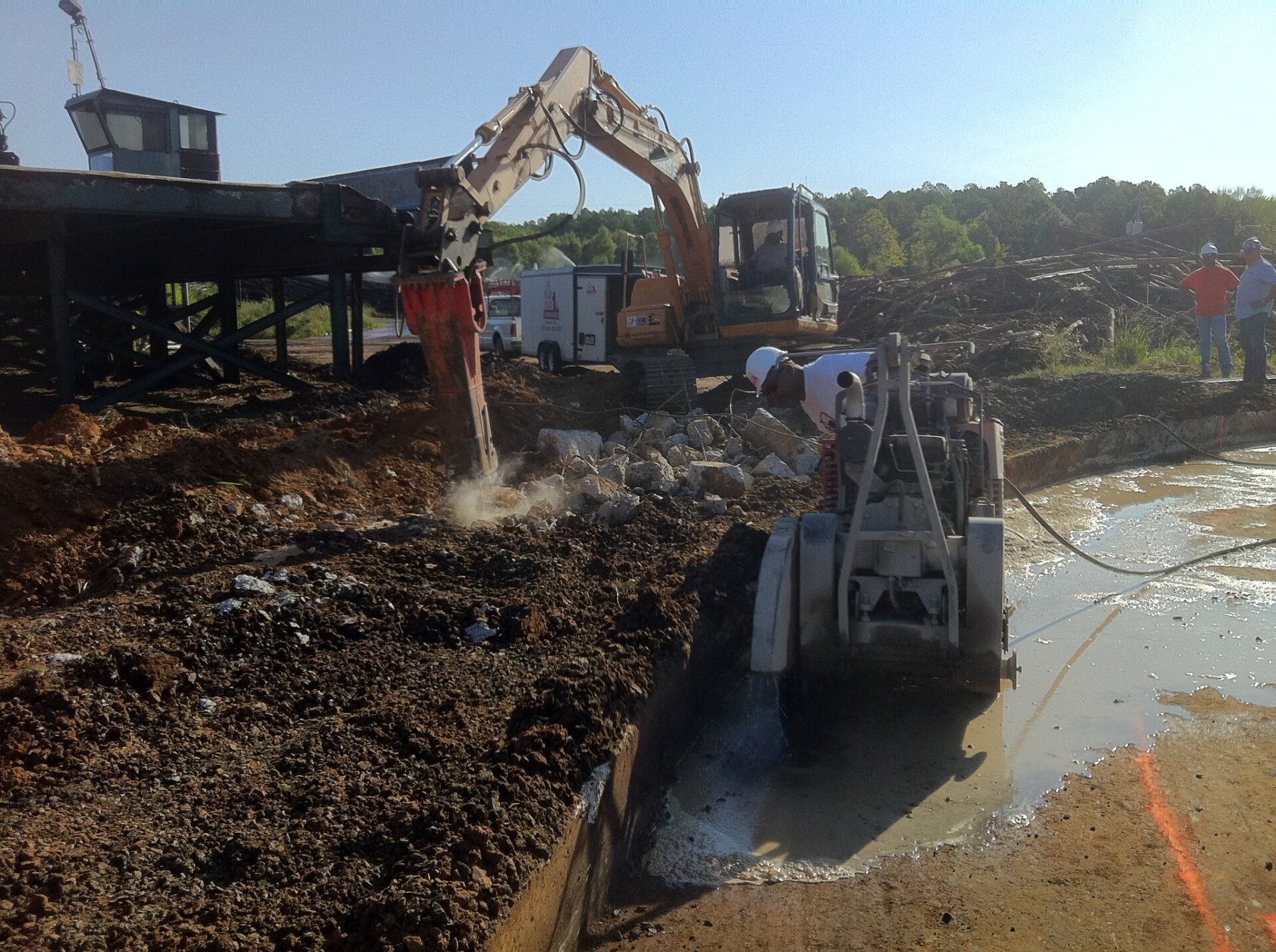Construction site with excavator and saw cutting through earth and debris, bridge in background.